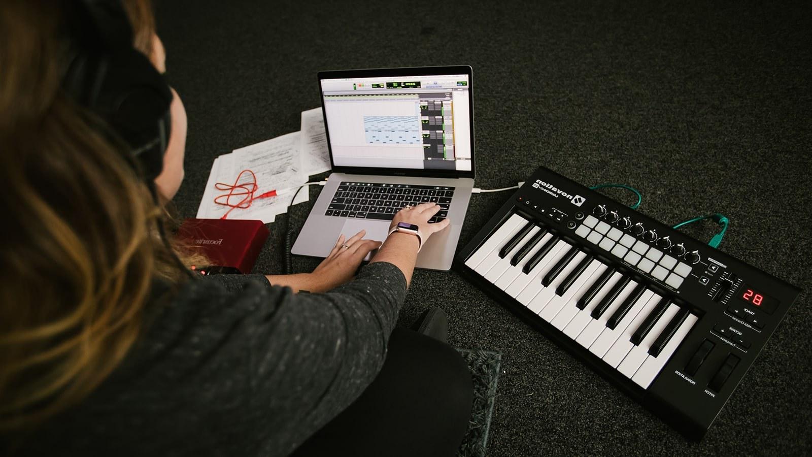 一个戴着耳机的学生坐在地板上，拿着笔记本电脑和电子键盘. Music editing software is on the laptop screen.