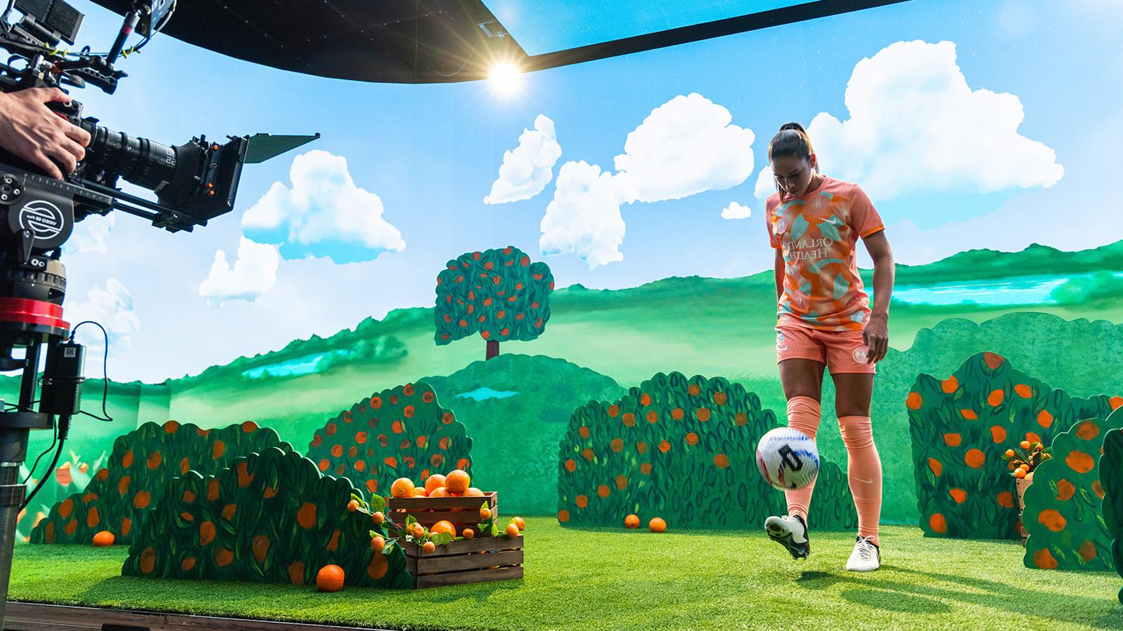 An Orlando Pride player kicks a soccer ball toward a camera on a film set that features an animated orange grove.