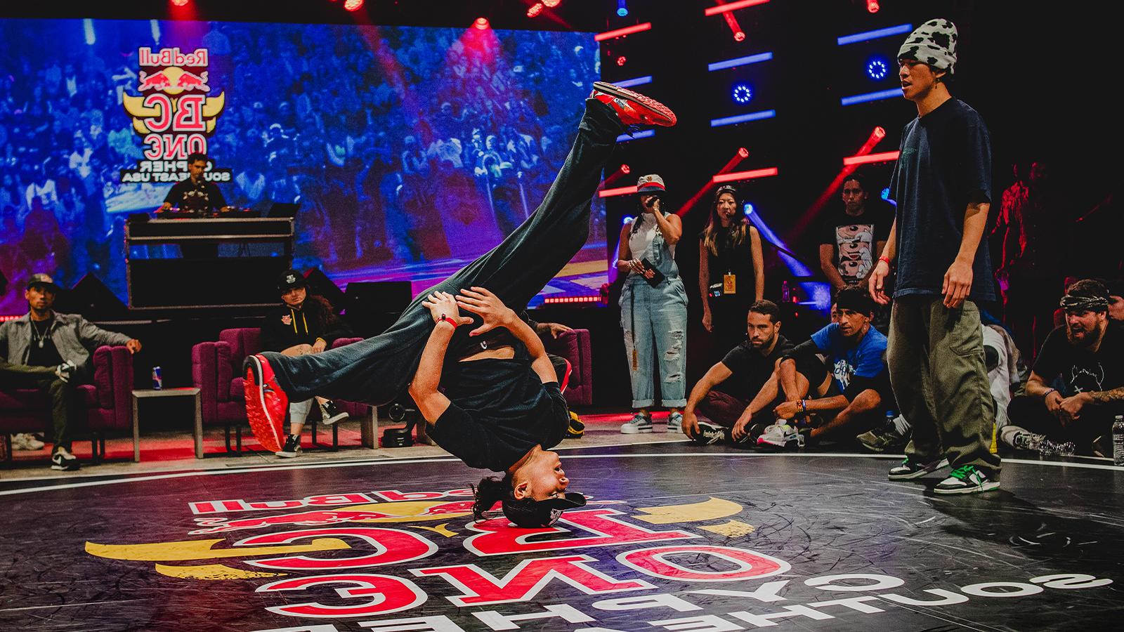 A dancer is doing a hands-free headstand with their legs spread while their competitor, 观众, and the judges watch.
