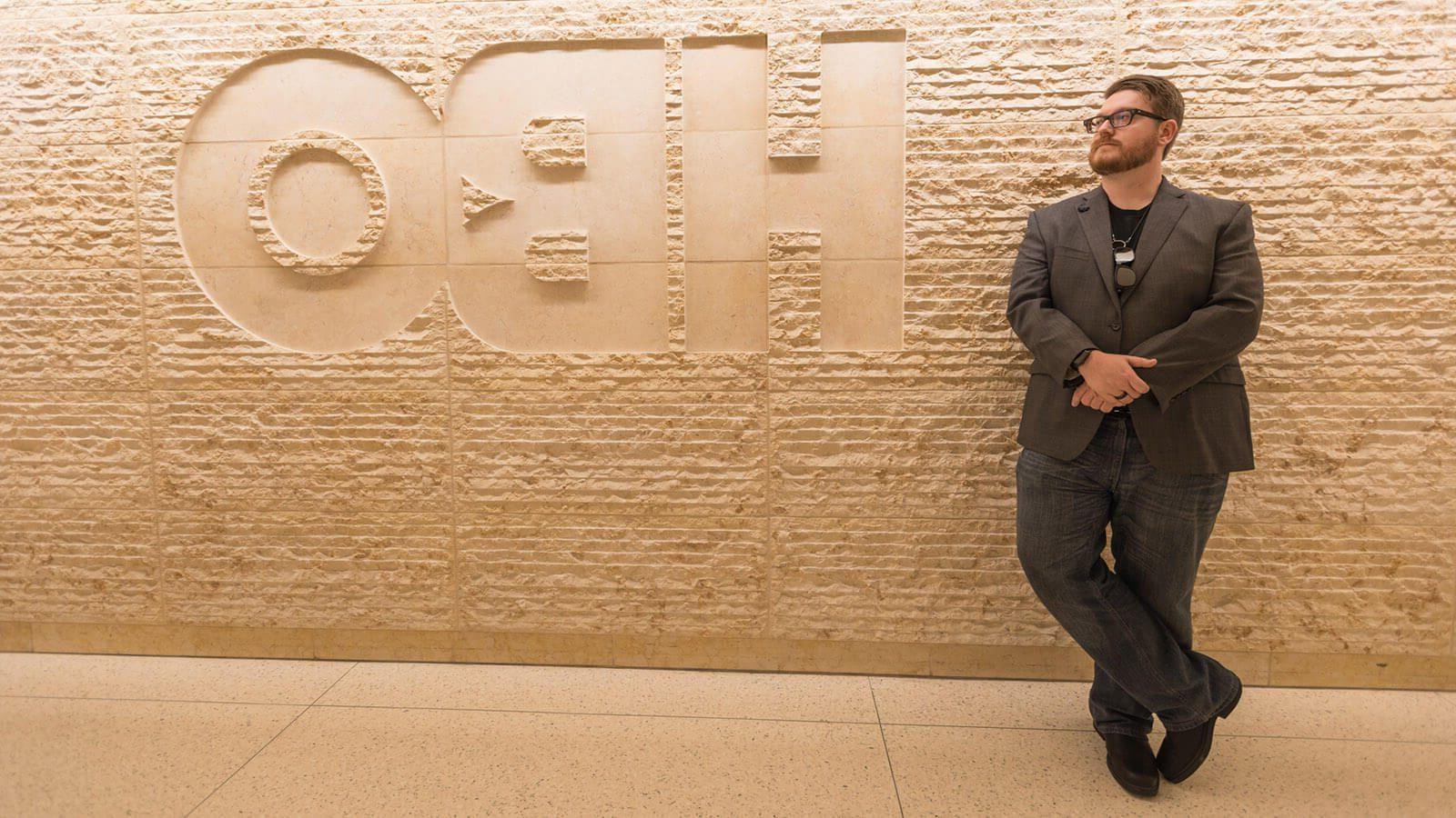 Wearing a suit and glasses, Stephen打转 leans against a beige, textured stone wall with the HBO logo carved into it.