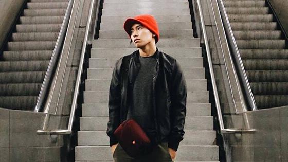 A young Asian man wearing a red knit cap, black jacket and shirt, green pants, and a burgundy bag stands between two escalators. 他双手插在口袋里，目光转向左边.