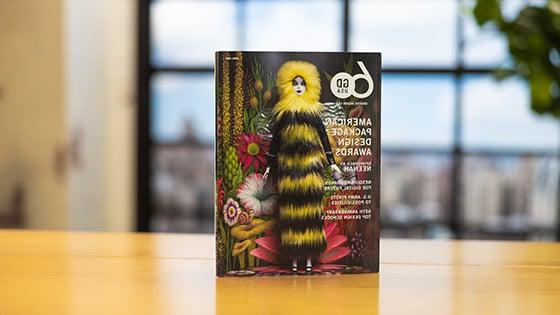 Graphic 设计 USA’s April 2023 issue sits upright on a wooden table. 的 cover features a female doll wearing a long furry black-and-yellow dress with a hood. She is standing in front of a psychedelic floral background.