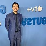 长得短的人, dark hair is smiling while wearing a blue suit against a light blue step and repeat that shows the logos for ‘Gutsy’ and Apple TV+.
