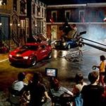 满帆 University's Backlot in the evening filled with a full film production crew shooting a scene featuring two muscle cars, one red 和 one black.
