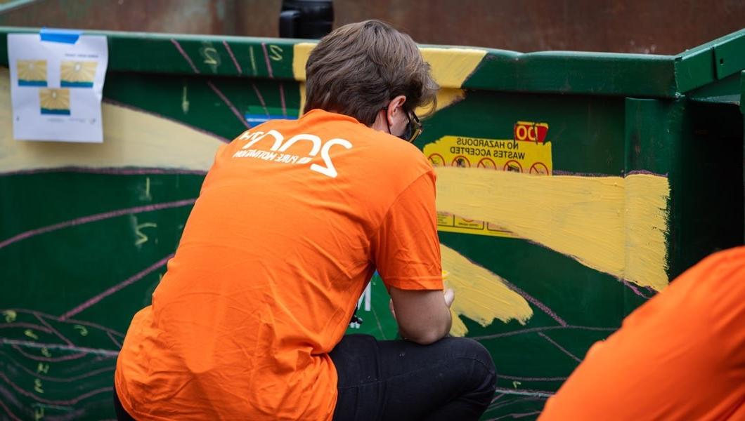 一名身穿亮橙色t恤的男子蹲下来，背对着镜头，正在给一个巨大的绿色垃圾桶作画.