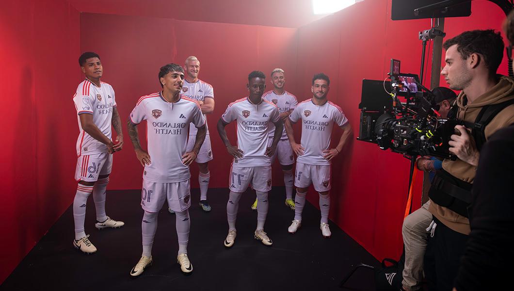 A group of six soccer players in full uniforms pose in a production studio while being filmed against a red backdrop.